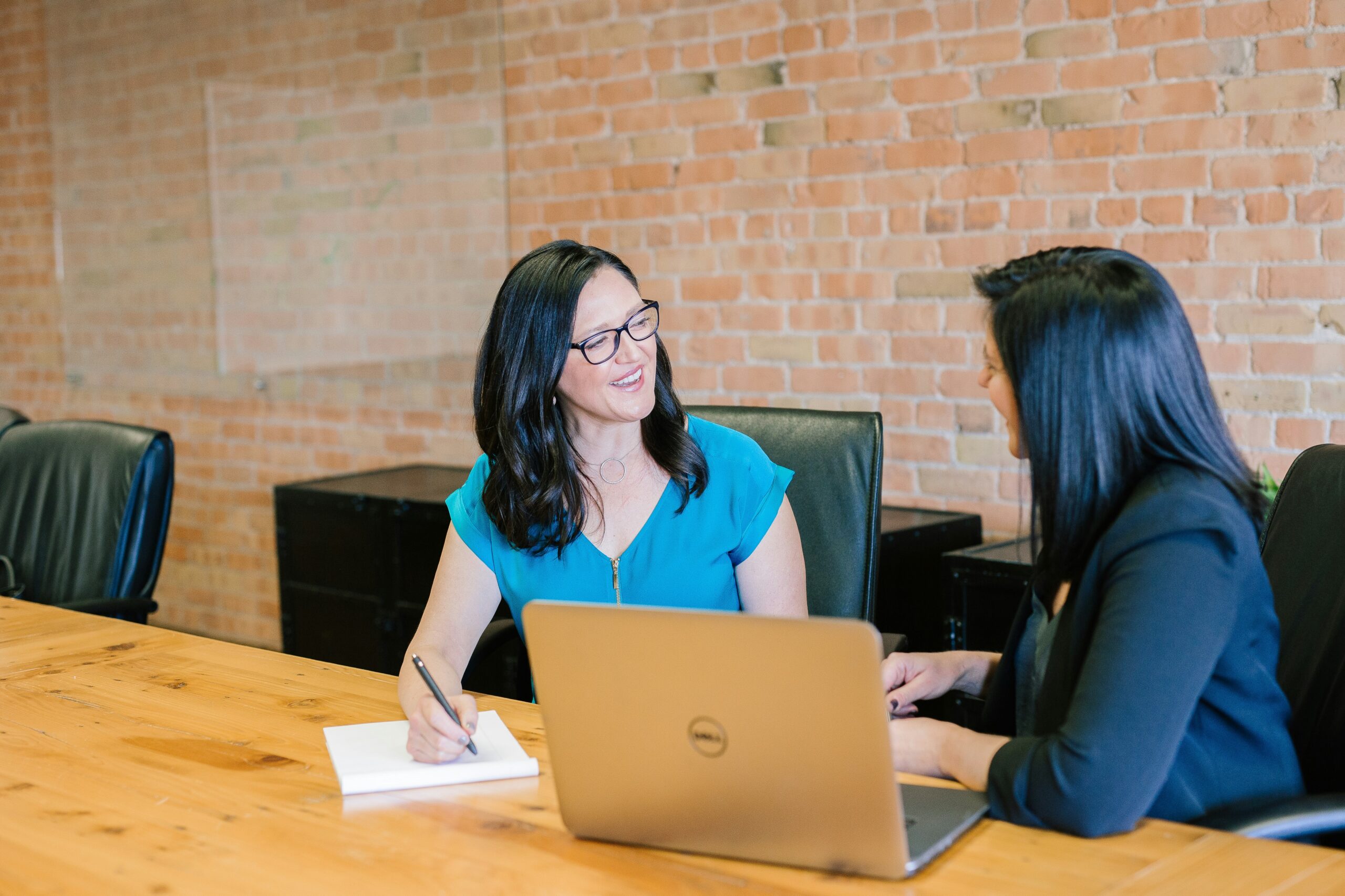 two people sitting in a conference room talking; one has a laptop and one has a notepad